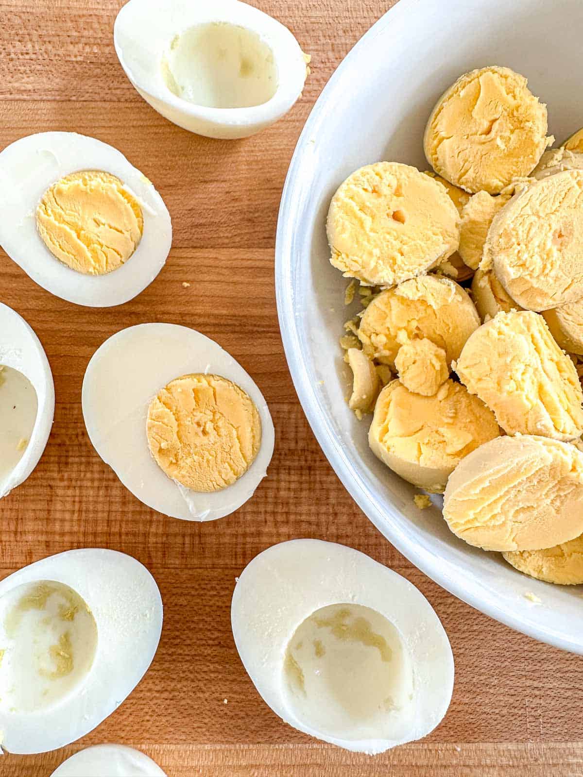 Halved hard-boiled eggs with yolks removed are on a wooden surface. The yolks are in a white bowl nearby, likely being prepared for deviled eggs.