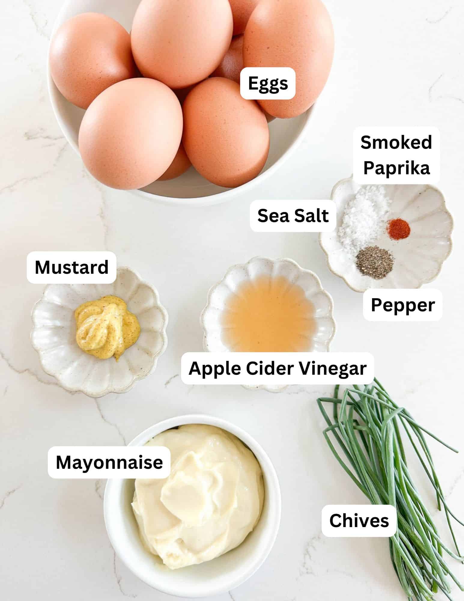 Bowl of eggs, a dish of mayonnaise, mustard, apple cider vinegar, fresh chives, and small plates with sea salt, pepper, and smoked paprika, all arranged on a white surface.