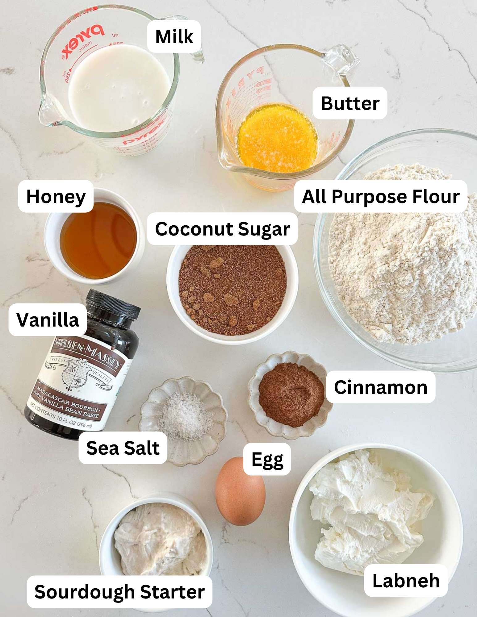 Ingredients for a recipe arranged on a white surface, labeled: milk, melted butter, honey, coconut sugar, flour, vanilla bean paste, sea salt, cinnamon, an egg, sourdough starter, and labneh.